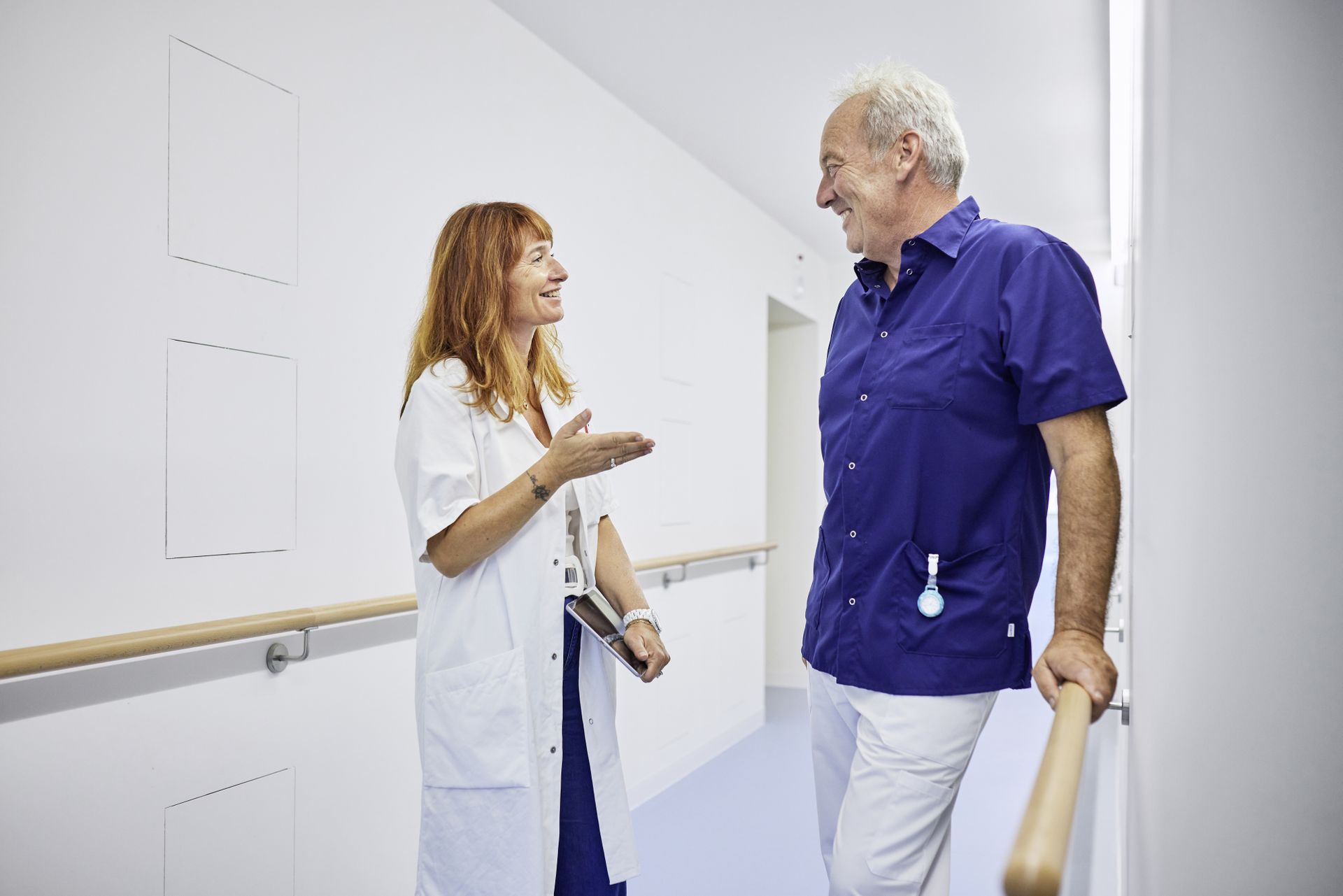 Mitarbeitende im Gesundheitswesen im persönlichen Austausch – Teamarbeit und Kommunikation sind zentrale Elemente im Pflegealltag. Zwei Pflegekräfte im Gespräch auf einem Klinikflur, eine davon mit Kittel, der andere in blauer Berufskleidung